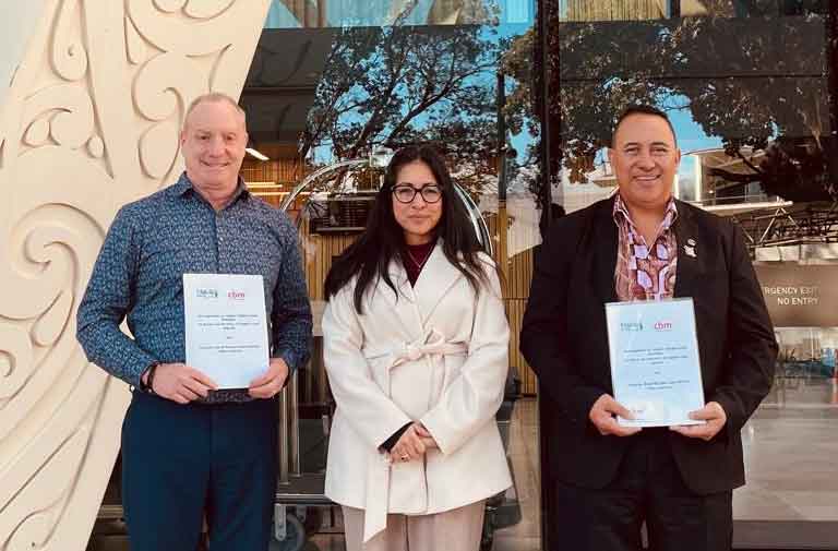 Three people stand outside a building with Māori carving, two of them holding signed copies of the partnership agreement.