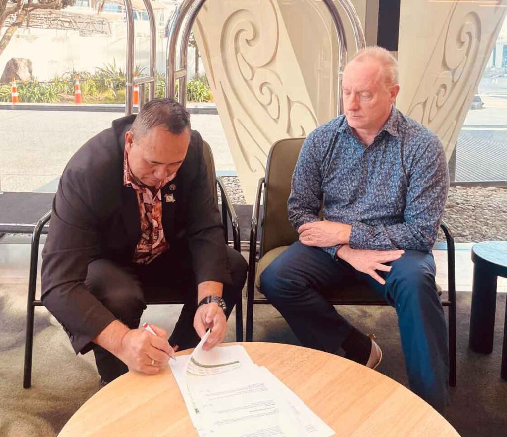 Two men seated at a table, one signing a copy of the partnership agreement while the other looks on.